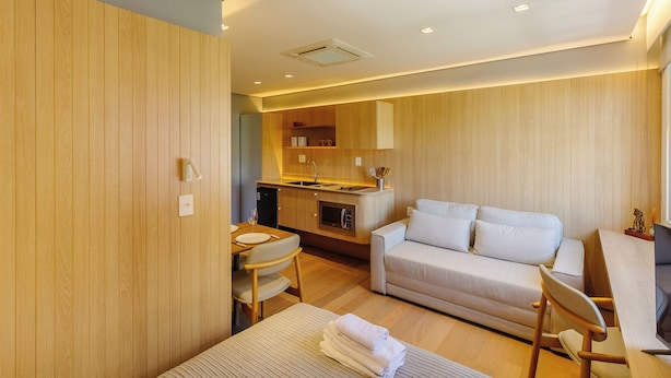 A modern hotel room with a wooden panel wall, a sofa, a dining table with chairs, and a kitchenette. - Beautiful Units in the Auxiliadora Neighborhood in PoA - CEN (Porto Alegre)