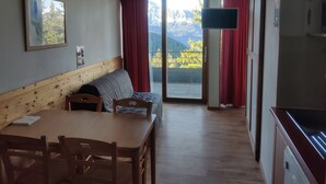 A room with a wooden dining table, chairs, a sofa, a TV, and a balcony with red curtains. - Apartment 6pers, balcony, mountain view (Chamrousse)