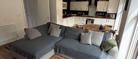 Un salon moderne avec un canapé d’angle gris, une table basse blanche et une cuisine en arrière-plan.