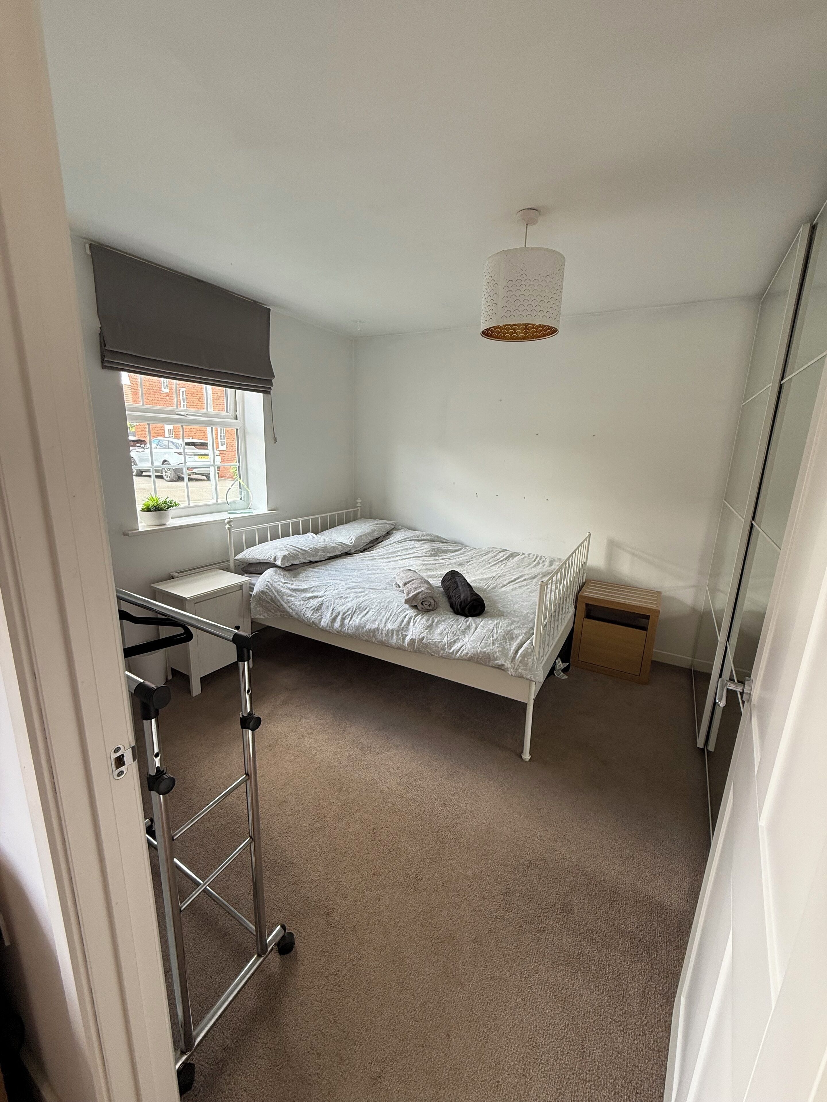 A bedroom with a single bed, a window with a view of a parking area, a bedside table, and a small wooden cabinet.