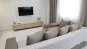 Un salon moderne doté d’un téléviseur à écran plat, d’un canapé beige, d’une console en bois et d’une grande fenêtre agrémentée de rideaux légers.