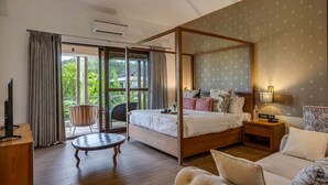 A bedroom with a four-poster bed, a small round table, a sofa, and a view of greenery through the glass doors.