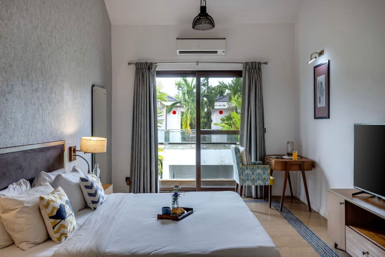 A hotel room with a bed, a desk, a chair, a TV, and a balcony with a view of palm trees.