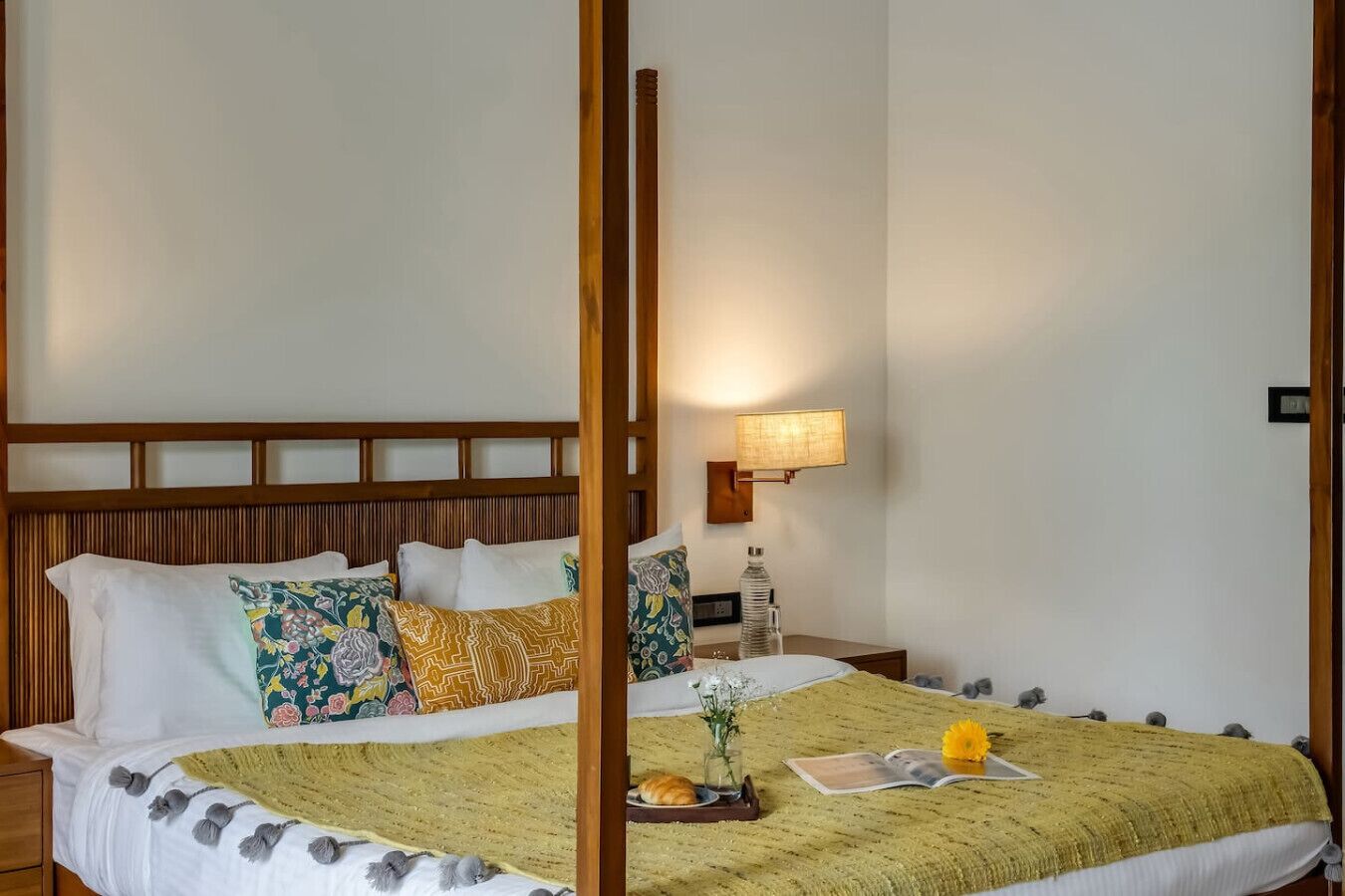 Een bed met een houten hoofdbord, meerdere kussens, een nachtkastje met een lamp en een decoratieve schaal met bloemen.