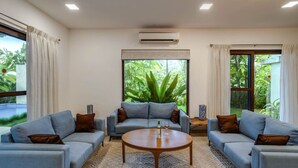 A modern living room with three sofas arranged around a central coffee table, large windows with curtains, and an air conditioning unit on the wall.