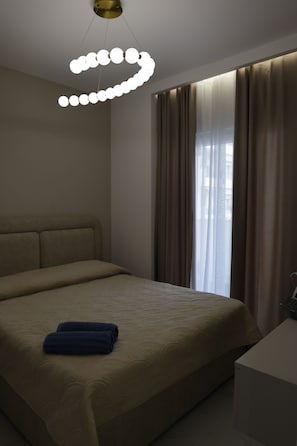 Ein Hotelzimmer mit Bett, Nachttisch, einem modernen Kronleuchter und einem Fenster mit Vorhängen.