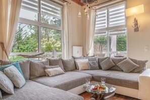 A cozy living room with a large sectional sofa, a glass coffee table with fruit, and a view of greenery outside. - Apartment 'Leuchturm' with Private Terrace, Private Garden and Wi-Fi (Rostock)