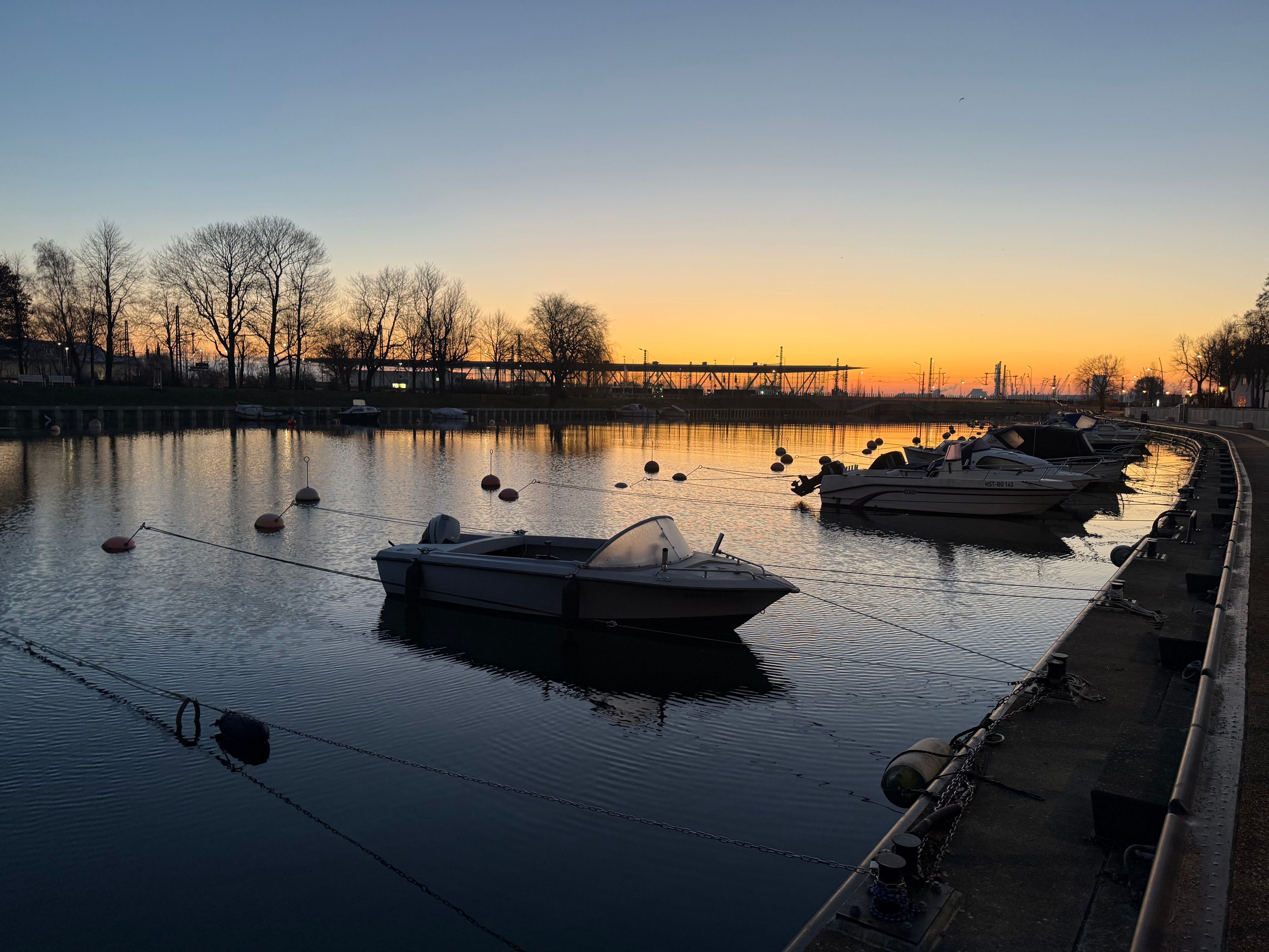 Een rustige rivier met aan de oever afgemeerde boten, een brug op de achtergrond en bomen langs de oevers.