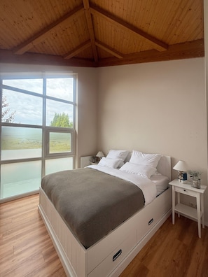 A bedroom with a large window, a bed with white bedding, a bedside table with a lamp, and a wooden ceiling.