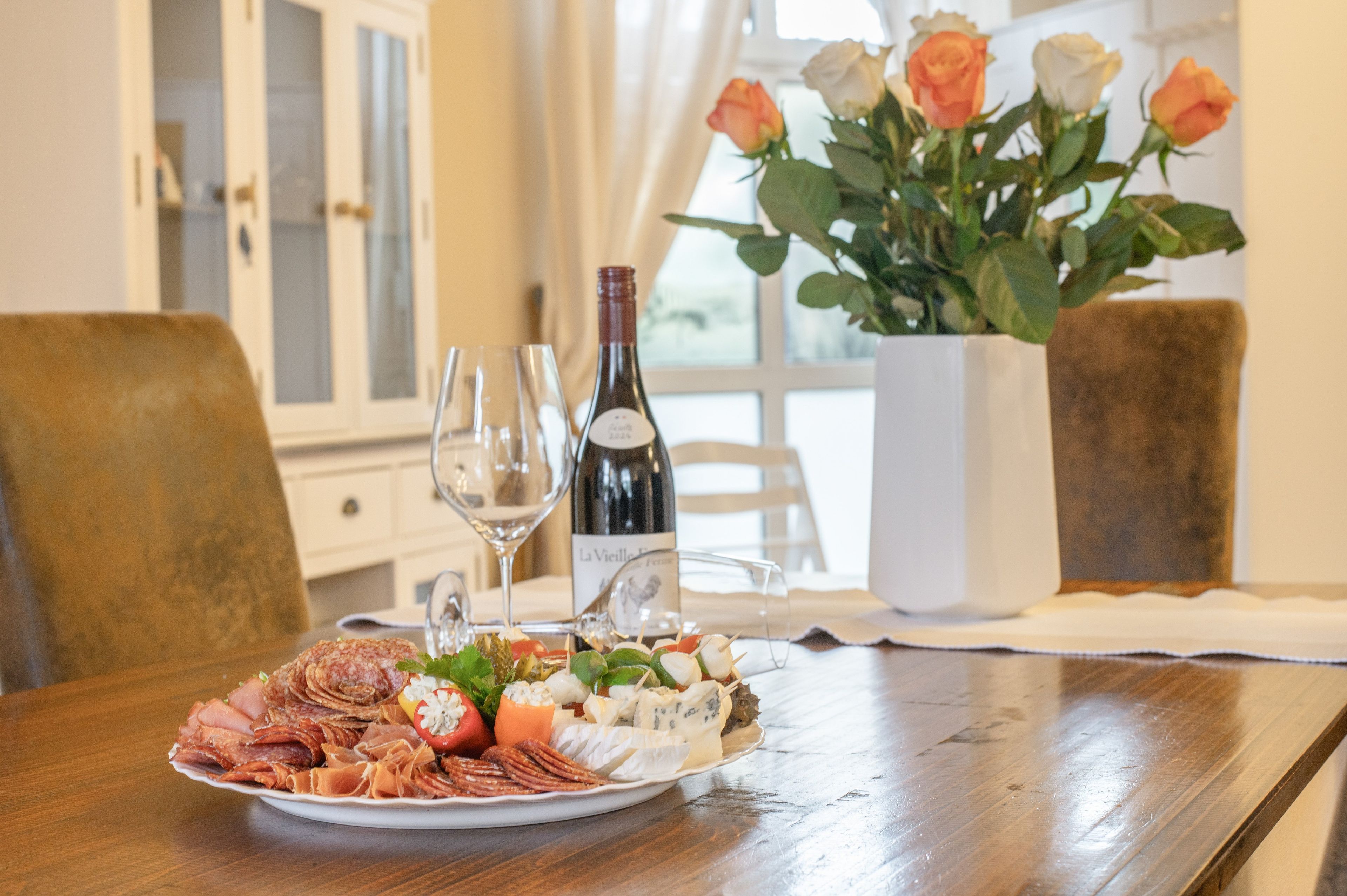 A dining table with a plate of assorted meats, cheeses, and vegetables, a bottle of wine, and two glasses. There are roses in a vase in the background.