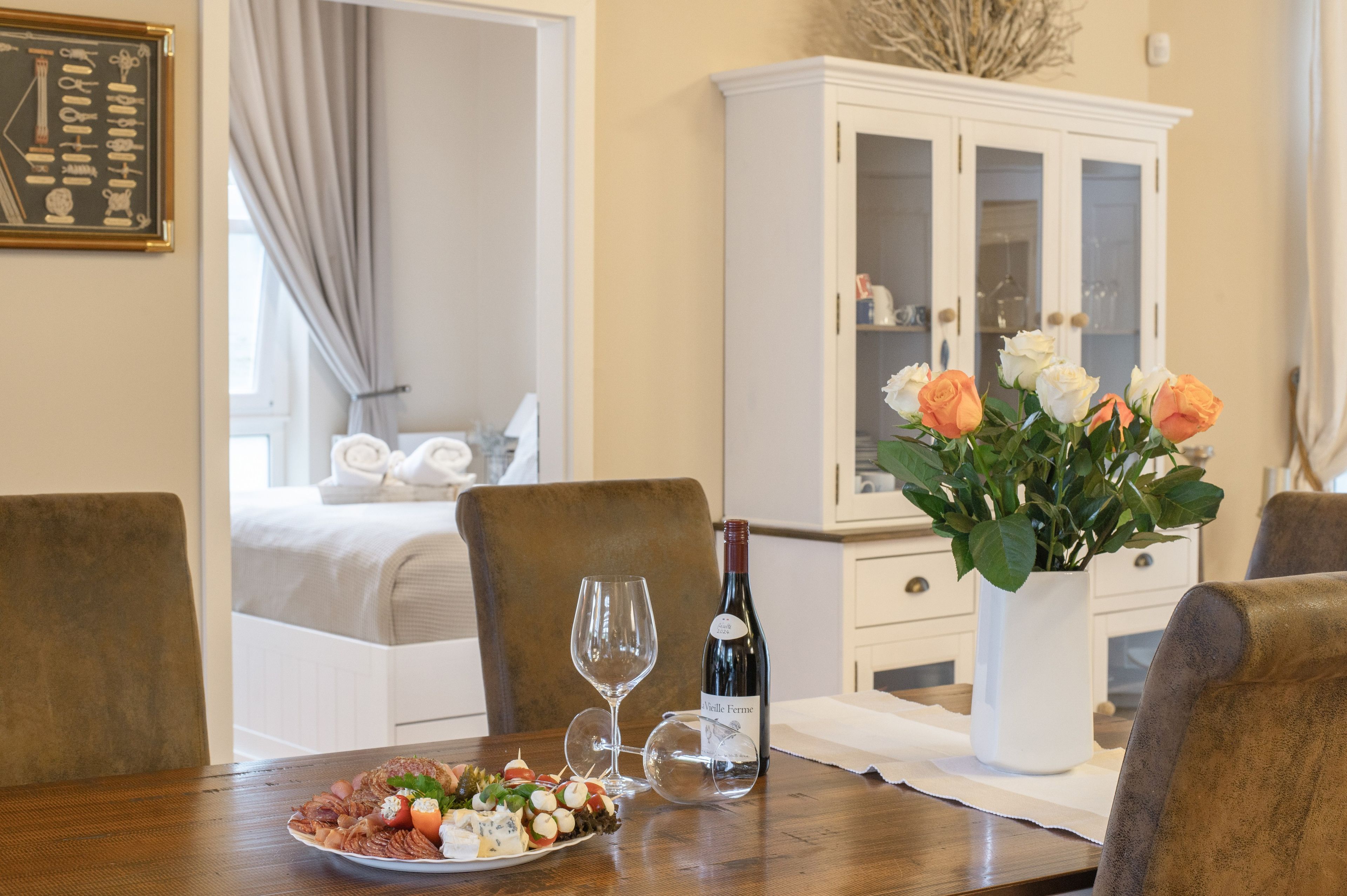 Un tavolo da pranzo con un piatto di antipasti, una bottiglia di vino e bicchieri, in una stanza con un letto e un armadio sullo sfondo.