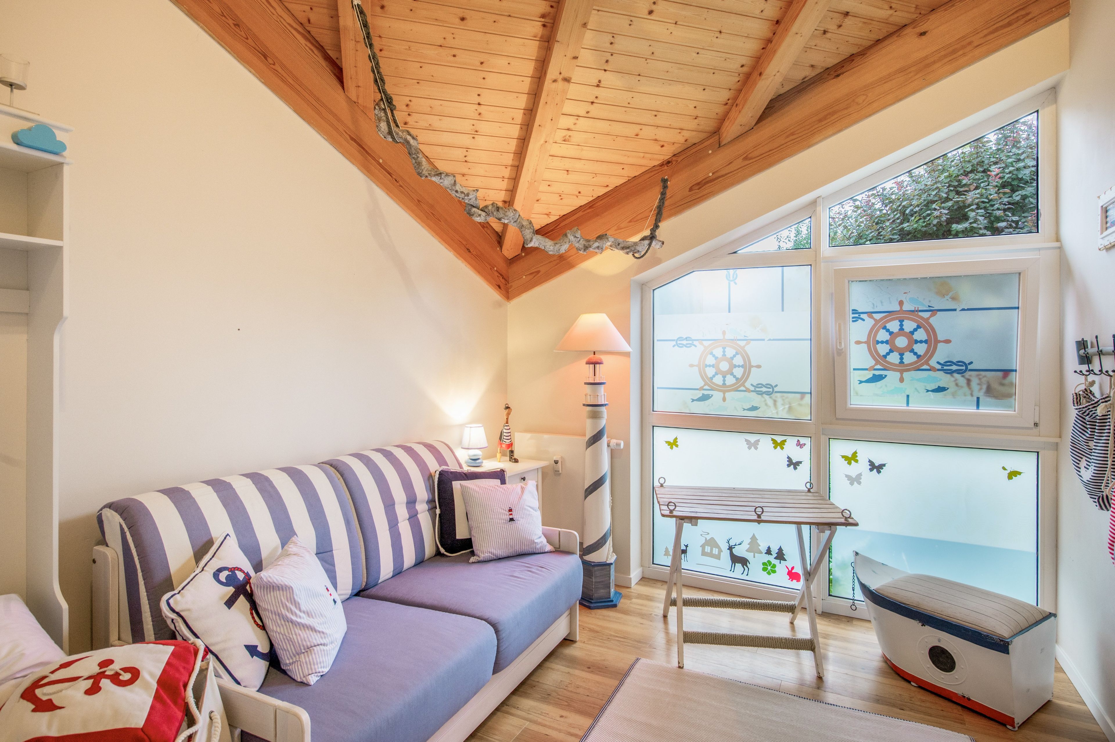 A cozy room with a wooden ceiling, a striped sofa, a small table, and a window with nautical-themed decorations.