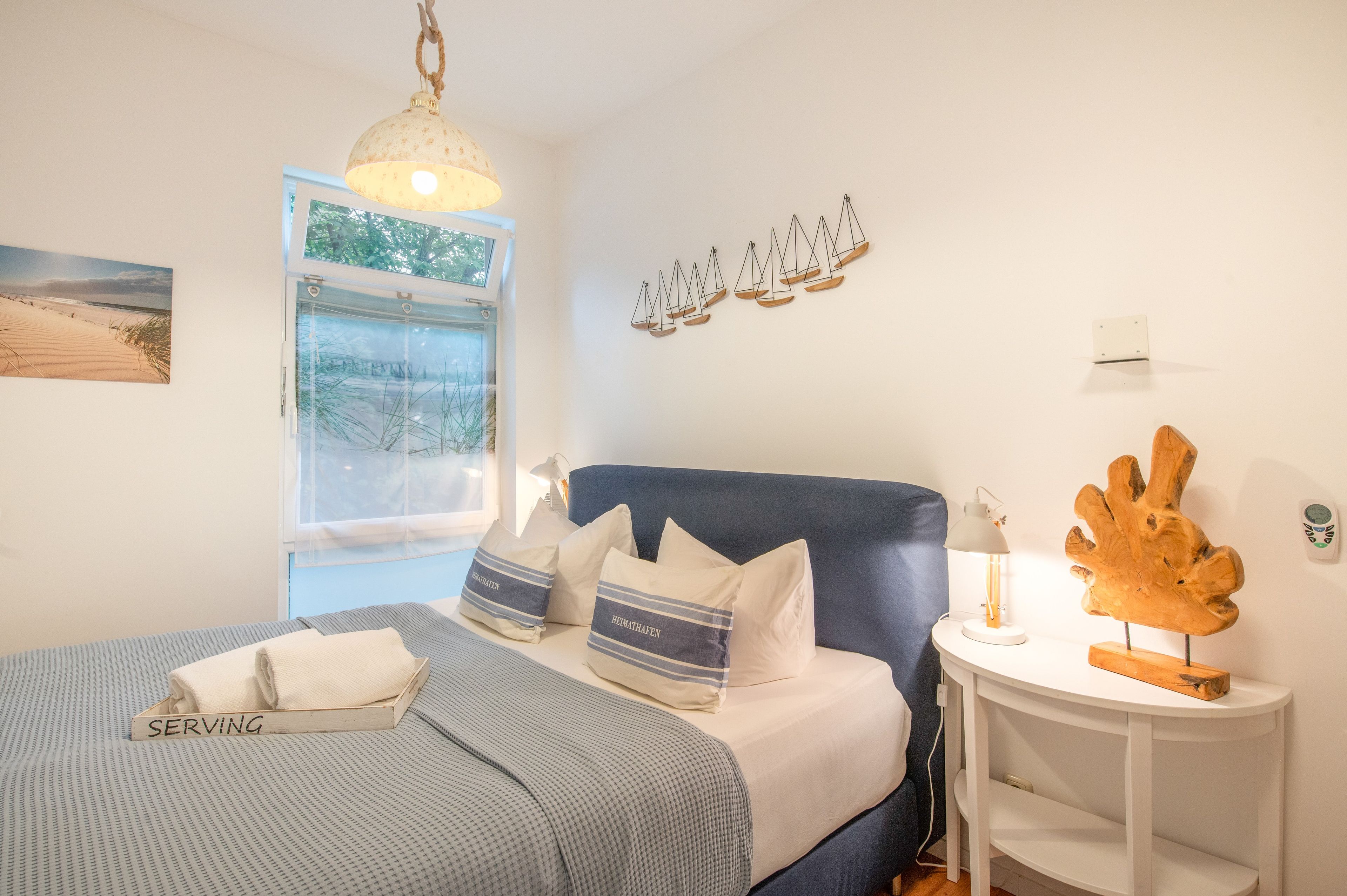 A bedroom with a bed, a window, a wall-mounted light fixture, and a small table with a lamp.
