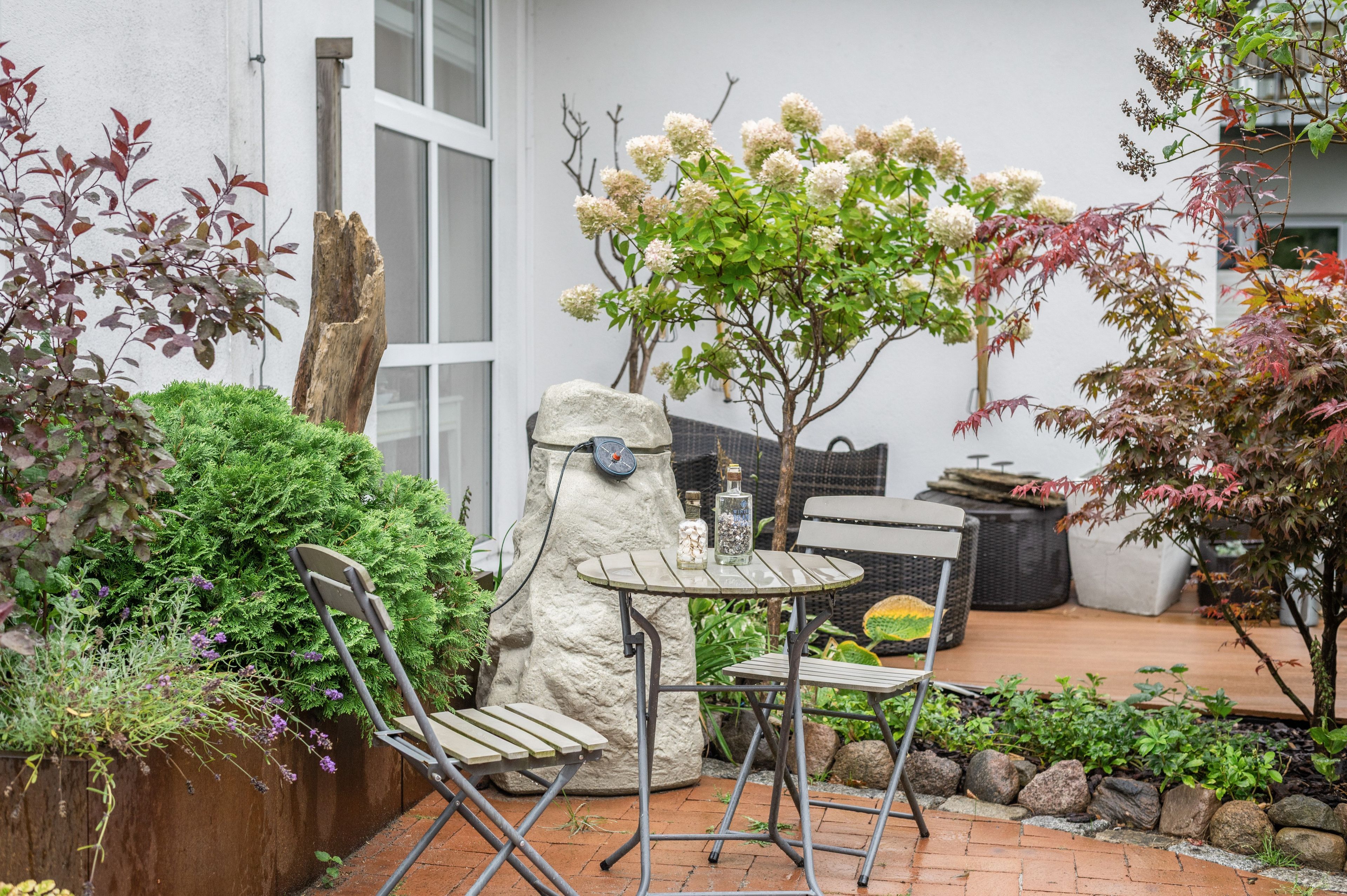 Eine Terrassenfläche mit Steintisch und zwei Stühlen, umgeben von Grünpflanzen und einem kleinen Baum mit weißen Blüten.