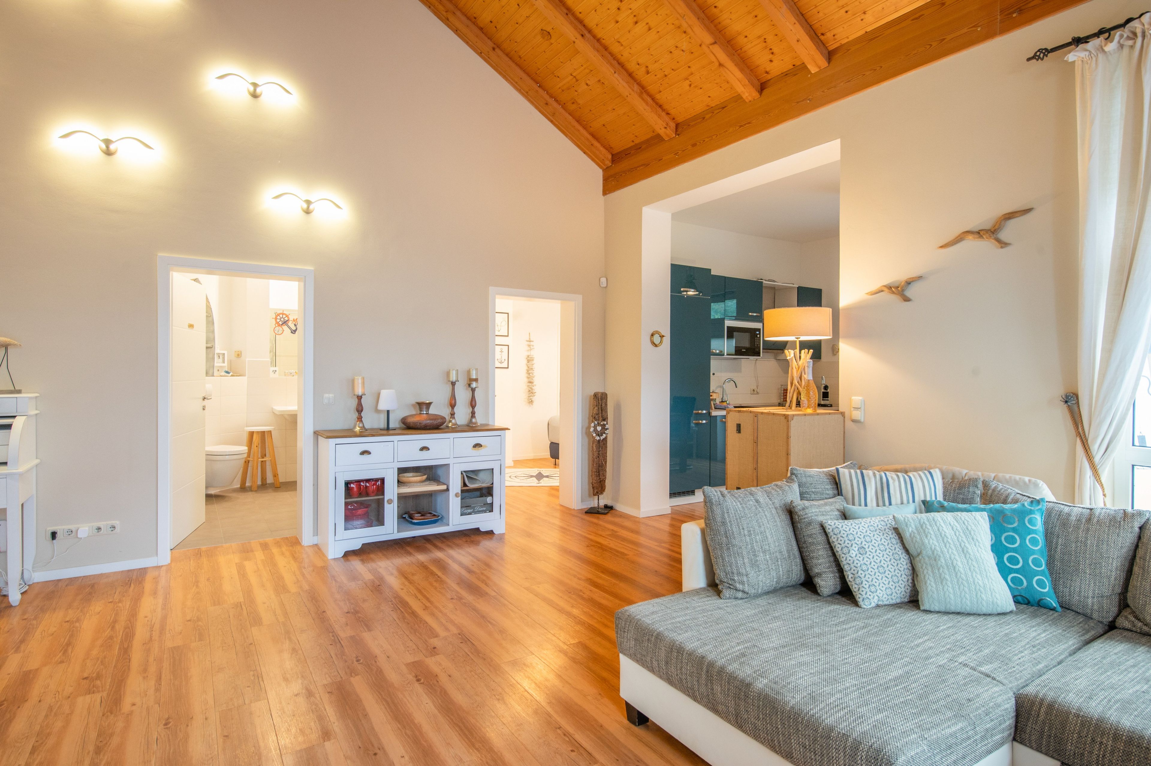 Un vaste espace de vie doté d’un plafond en bois, d’un canapé agrémenté de coussins à motifs et d’une cuisine visible à travers une porte ouverte.