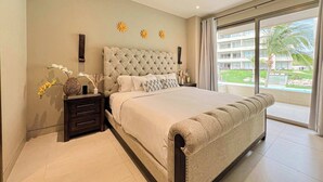 A bedroom with a large bed, a ceiling fan, a nightstand, and a window with a view of a building and greenery. - Vista Vela Condo · Pool, Gym & Pickleball Courts (El tezal)