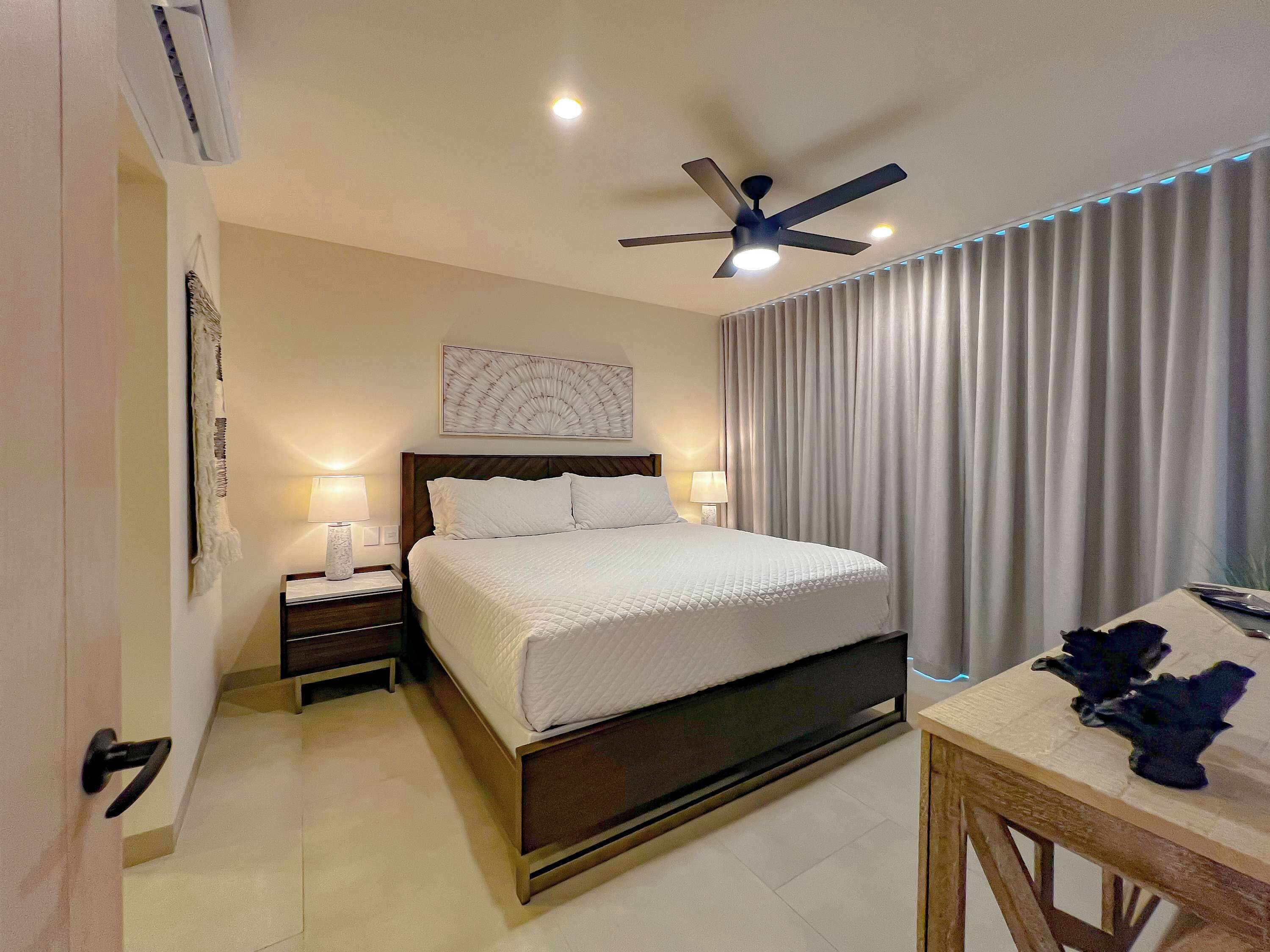 A hotel room with a bed, bedside tables, a ceiling fan, and a large window with curtains.