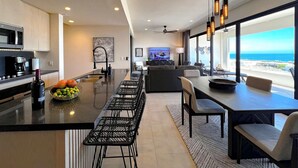 A modern kitchen with a central island, bar stools, and a dining area with a view of the ocean. - Private Rooftop Terrace, Ocean Views & Pool Access (Cabo San Lucas)