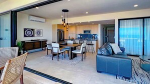 A modern living room with a sofa, dining table, and chairs. There is a kitchen area with a microwave and refrigerator. The room has large windows and a view of the outside. - Private Rooftop Terrace, Ocean Views & Pool Access (Cabo San Lucas)