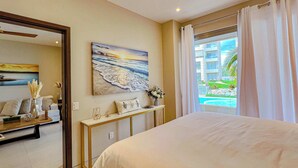 Ein Schlafzimmer mit einem großen Bett, einem Bild einer Strandansicht, einem Konsolentisch mit dekorativen Gegenständen und Blick auf einen Swimmingpool und Gebäude.