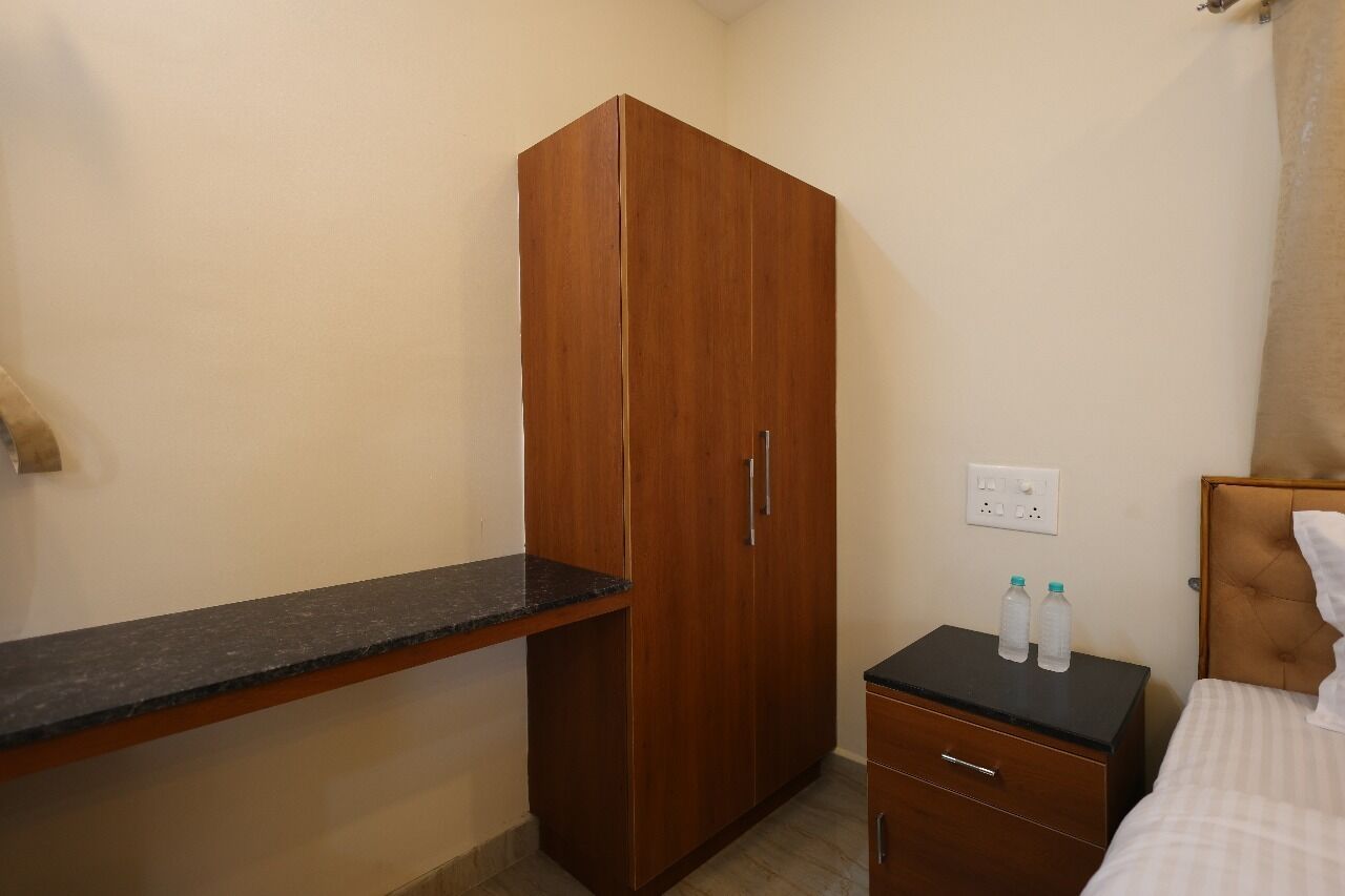 A bedroom with a wooden wardrobe, a small bedside table with two bottles, a bed with a headboard, and a nightstand.