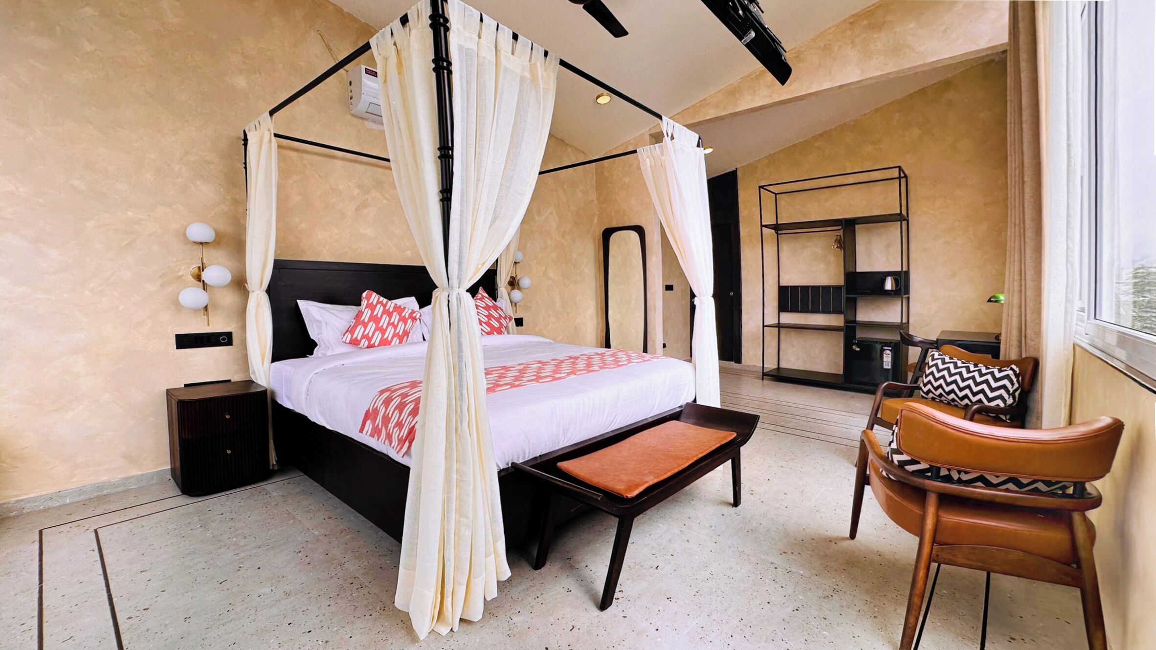 A four-poster bed with a canopy, a bedside table, a chair, and a shelving unit in a hotel room.