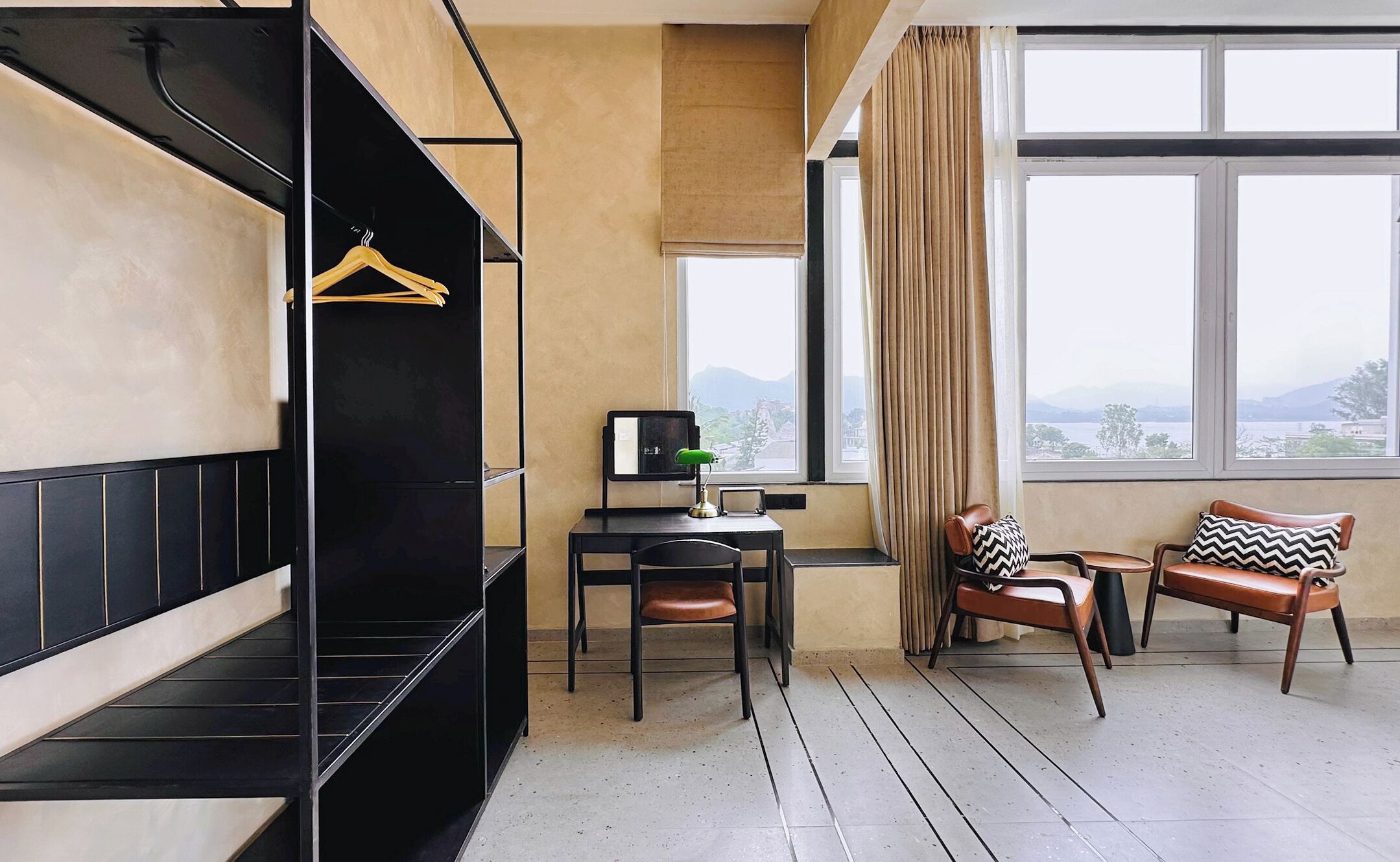 A room with a black sofa, a desk with a lamp, two chairs, a window with a view, and a hanging coat rack.
