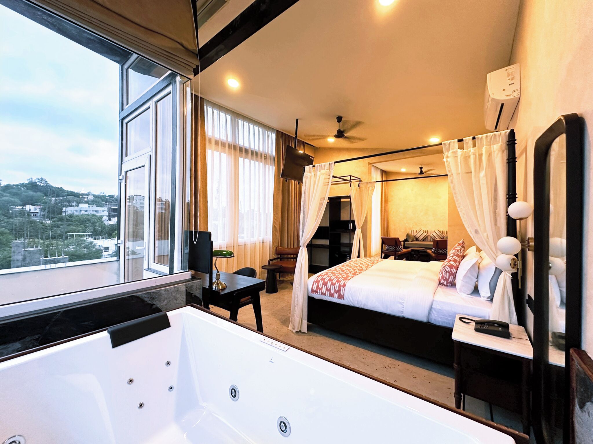 A hotel room with a large bed, a sitting area with a table and chairs, a jacuzzi, and a view of the city.