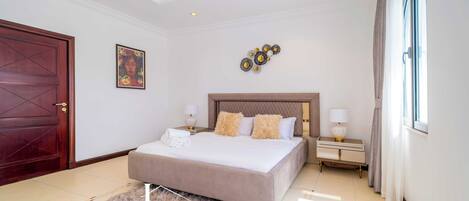 A spacious bedroom with a large bed, a wooden door, a painting, and a decorative wall piece.