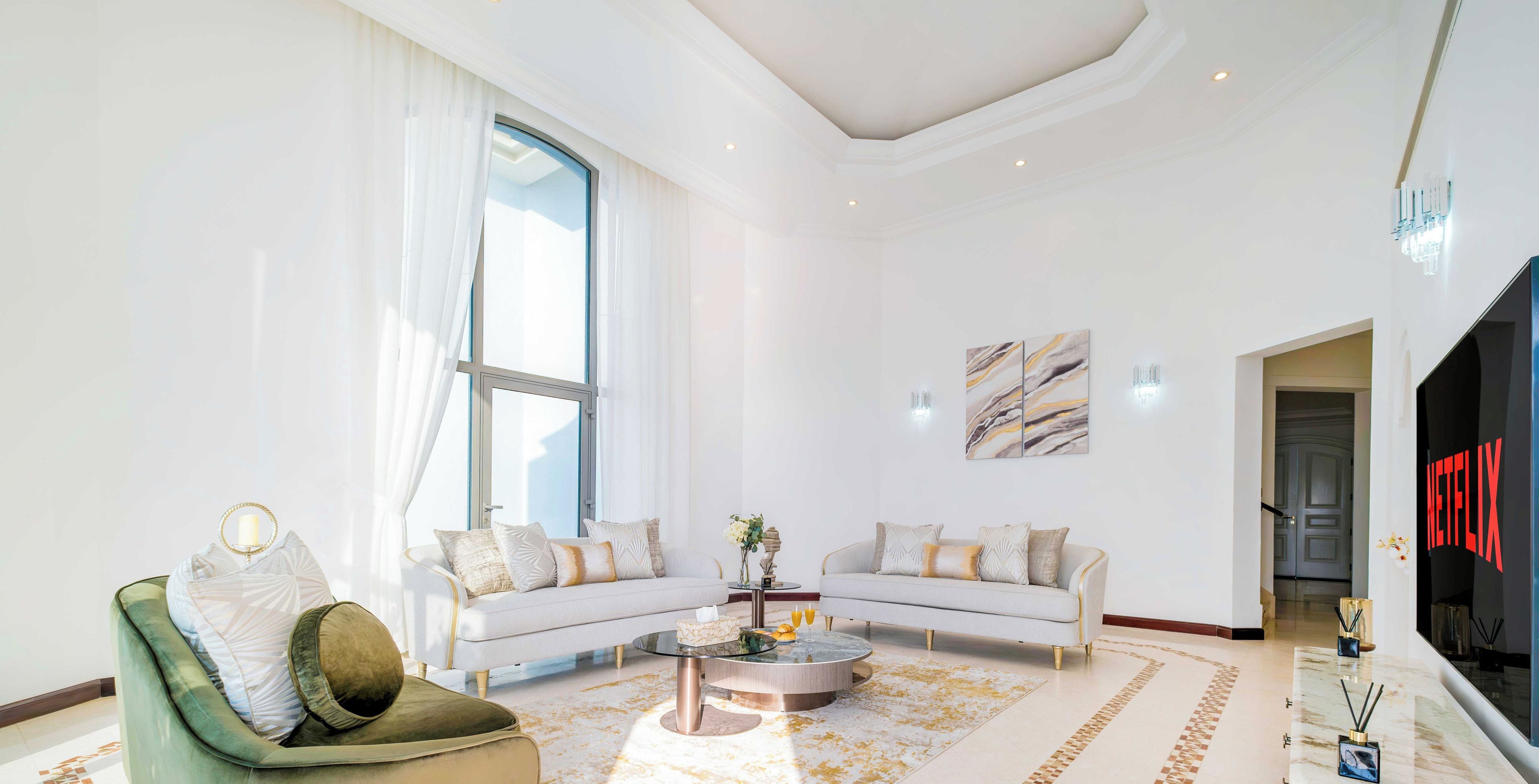 Una sala de estar moderna con un sillón verde, un sofá blanco y una mesa de centro. Hay un ventanal con cortinas blancas, un televisor de pantalla plana y una alfombra decorativa.