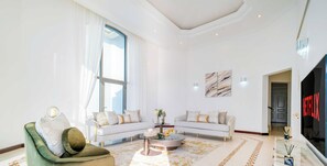 Een moderne woonkamer met een groene fauteuil, een witte bank en een salontafel. Er is een groot raam met witte gordijnen, een flatscreen televisie en een decoratief vloerkleed.