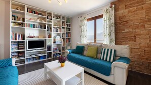 Un salon comprenant un canapé, une table basse, une bibliothèque, un téléviseur et une fenêtre avec des rideaux.