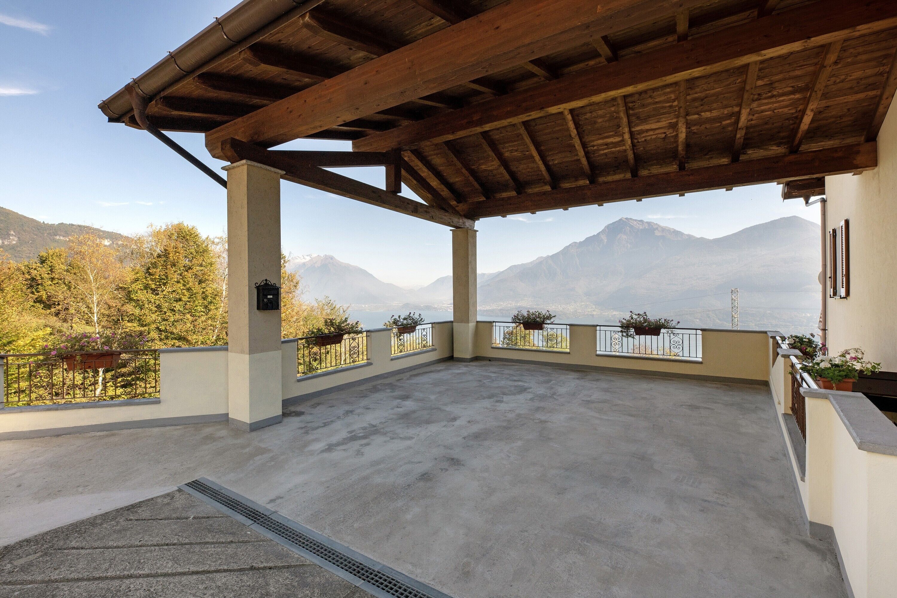 A covered outdoor terrace with a view of mountains, a concrete floor, and potted plants along the railing.