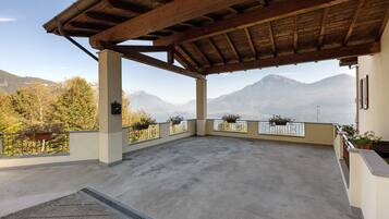 A covered outdoor terrace with a view of mountains, a concrete floor, and potted plants along the railing.
