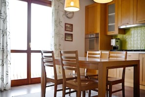 Uma sala de jantar com cadeiras e mesa de madeira, uma cozinha com armários de madeira, um relógio na parede e uma janela com cortinas.