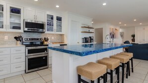 Una cocina moderna con armarios blancos, una isla de mármol azul, electrodomésticos de acero inoxidable y una zona de estar con taburetes de mimbre.