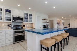 Una cocina moderna con armarios blancos, una isla de mármol azul, electrodomésticos de acero inoxidable y una zona de estar con taburetes de mimbre.