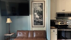 Una sala de estar con un sofá marrón, una mesa de centro de madera, un tapete estampado, un televisor de pantalla plana, un adorno en la pared y una cocina con electrodomésticos modernos.