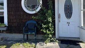 Una casa de color marrón oscuro, con una puerta blanca con el número 74, un ventanal redondo con un diseño decorativo de vidrio y una silla de plástico azul en el exterior.