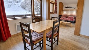 A dining area with a wooden table and chairs, a view of a snowy landscape through large windows, and a cozy seating area with patterned cushions. - 10m from ski slopes, 2nd floor, balcony, ski locker, 36m², Méribel-Mottaret (Méribel - Les Allues)