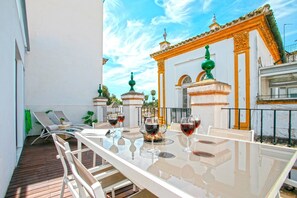 Un espacio al aire libre con mesas y sillas blancas, junto a una pared blanca y un arco decorativo bañado por el sol.
