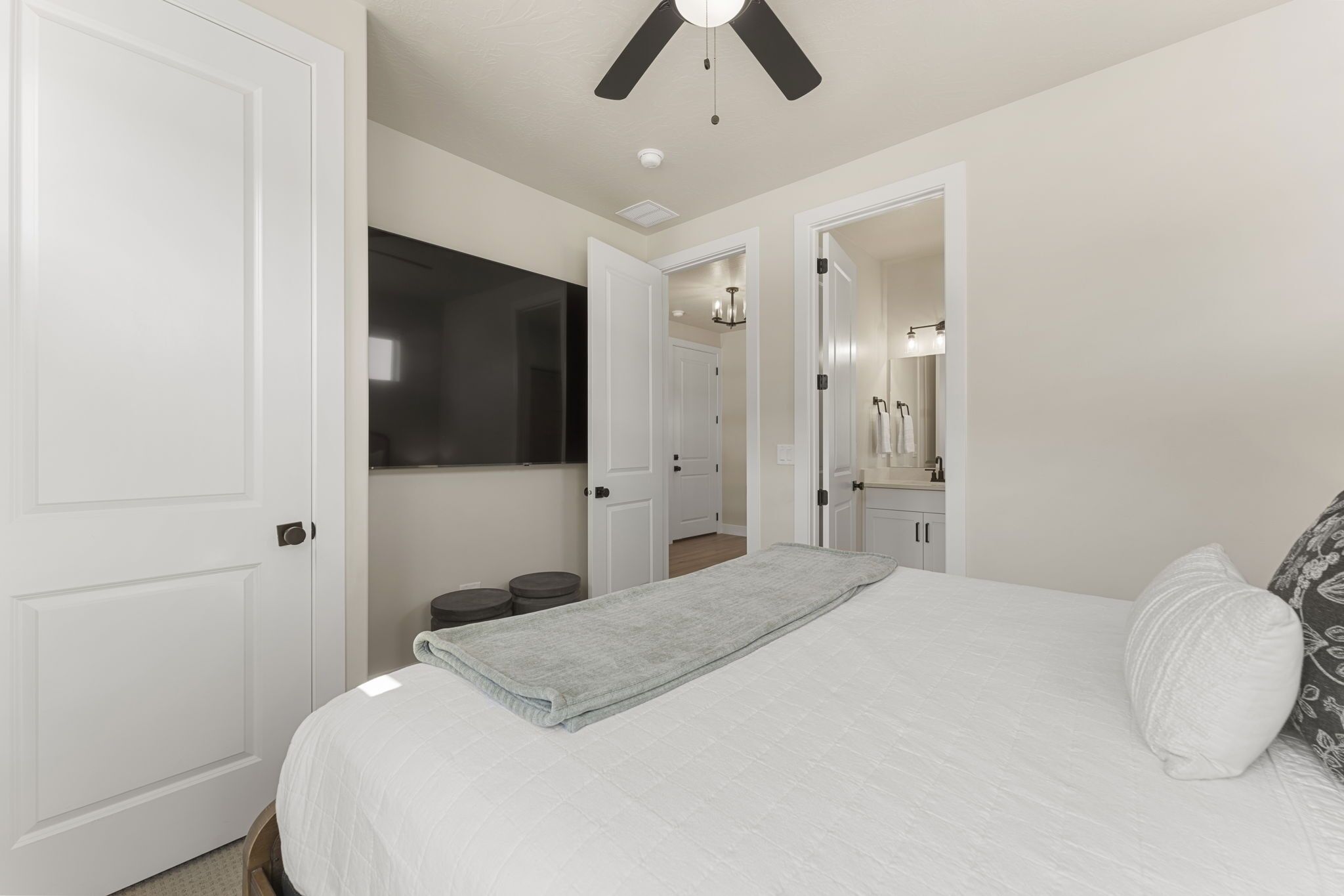 Un dormitorio con cama, ventilador de techo, una puerta blanca y un pasillo que lleva a un baño.