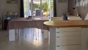 Et moderne køkken med en central ø, et spiseområde med bord og stole, samt udsigt til naturen gennem store vinduer.