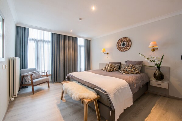 A modern bedroom with a large bed, a chair, a bench, and a nightstand with a lamp. - Inn Bruges Suites (Bruges)