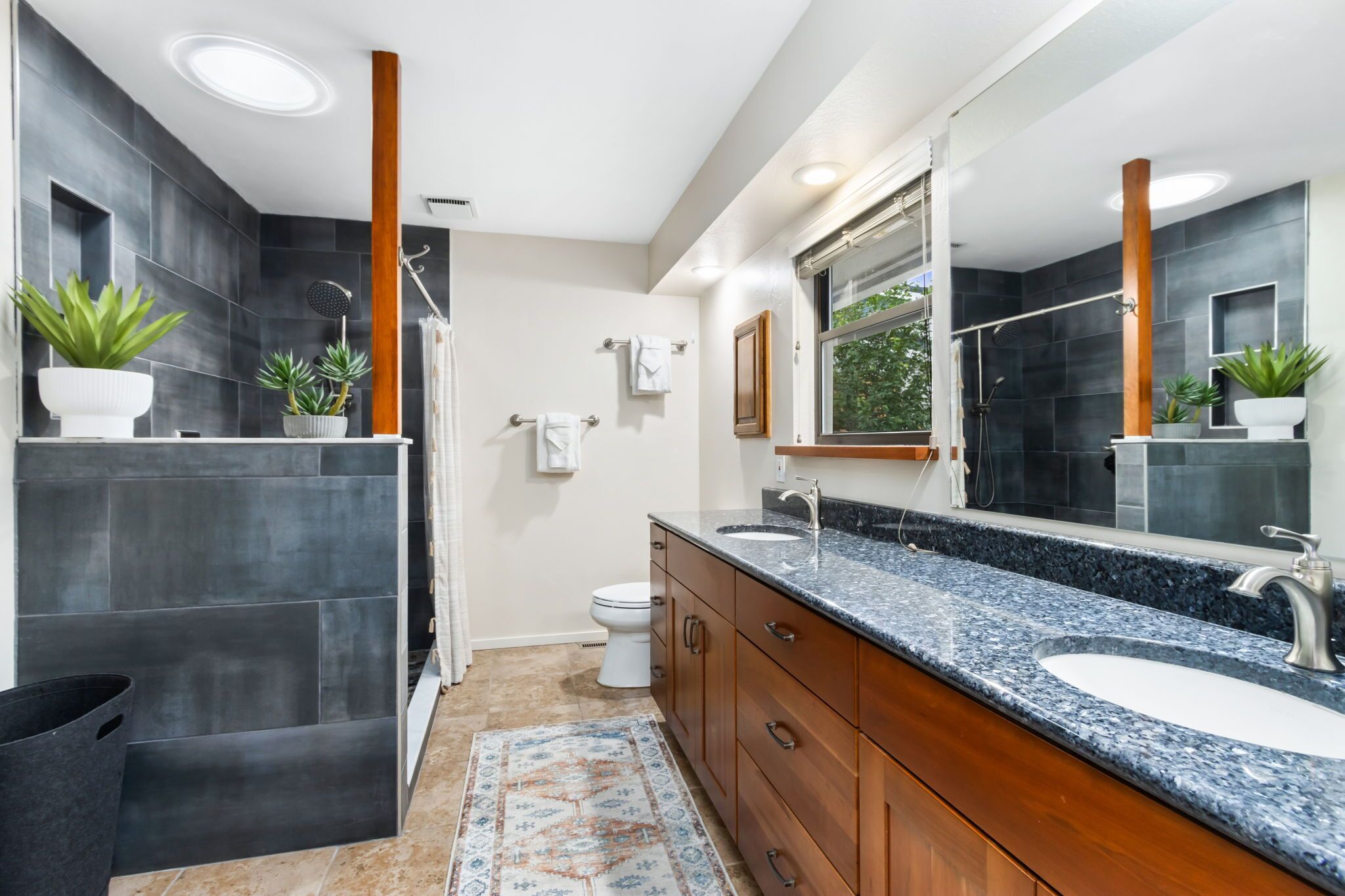 A modern bathroom with dark tiles, a large mirror, a wooden vanity, and a window.