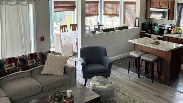 A living room with a sofa, armchair, coffee table, and a rug. There is a kitchen area with a microwave and cabinets. - Fabulous 3-bedroom Weirs Beach Village home in Laconia. (Laconia)
