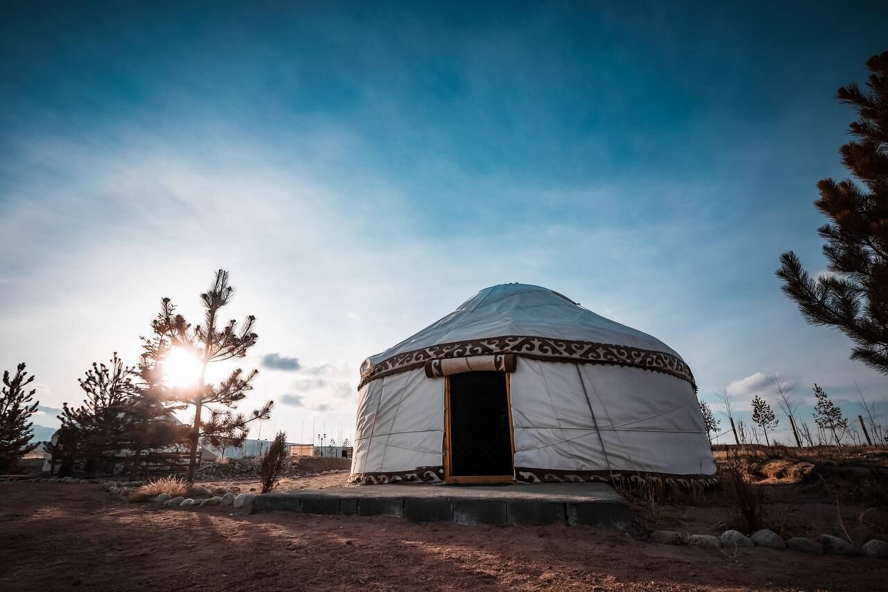 A traditional yurt with a circular structure and a door, surrounded by sparse vegetation and a clear sky.