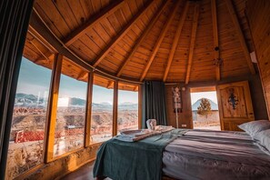 A wooden room with a bed, large windows, and a view of a mountainous landscape.