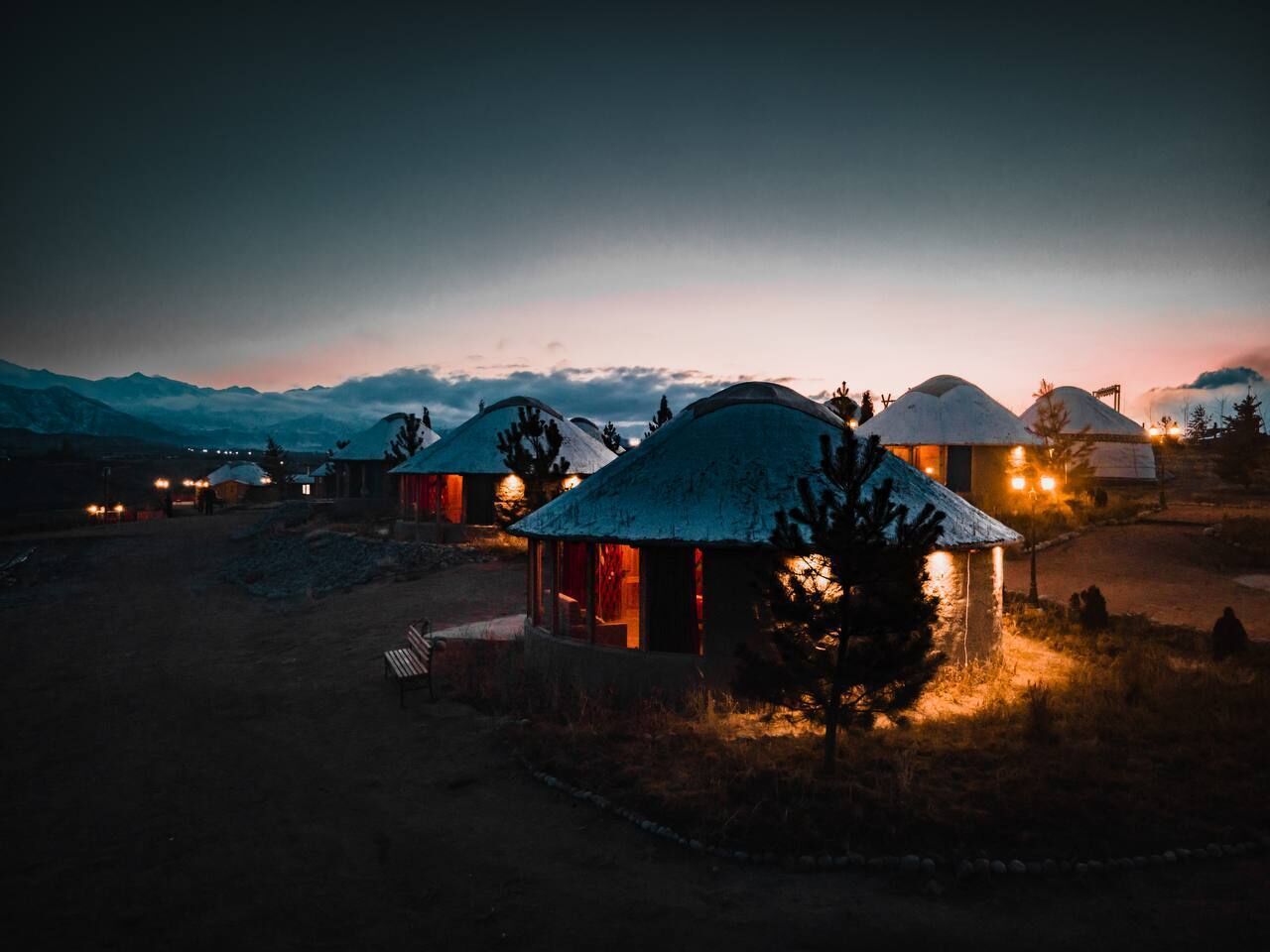 A series of dome-shaped cabins with warm interior lighting, set against a mountainous backdrop during twilight.