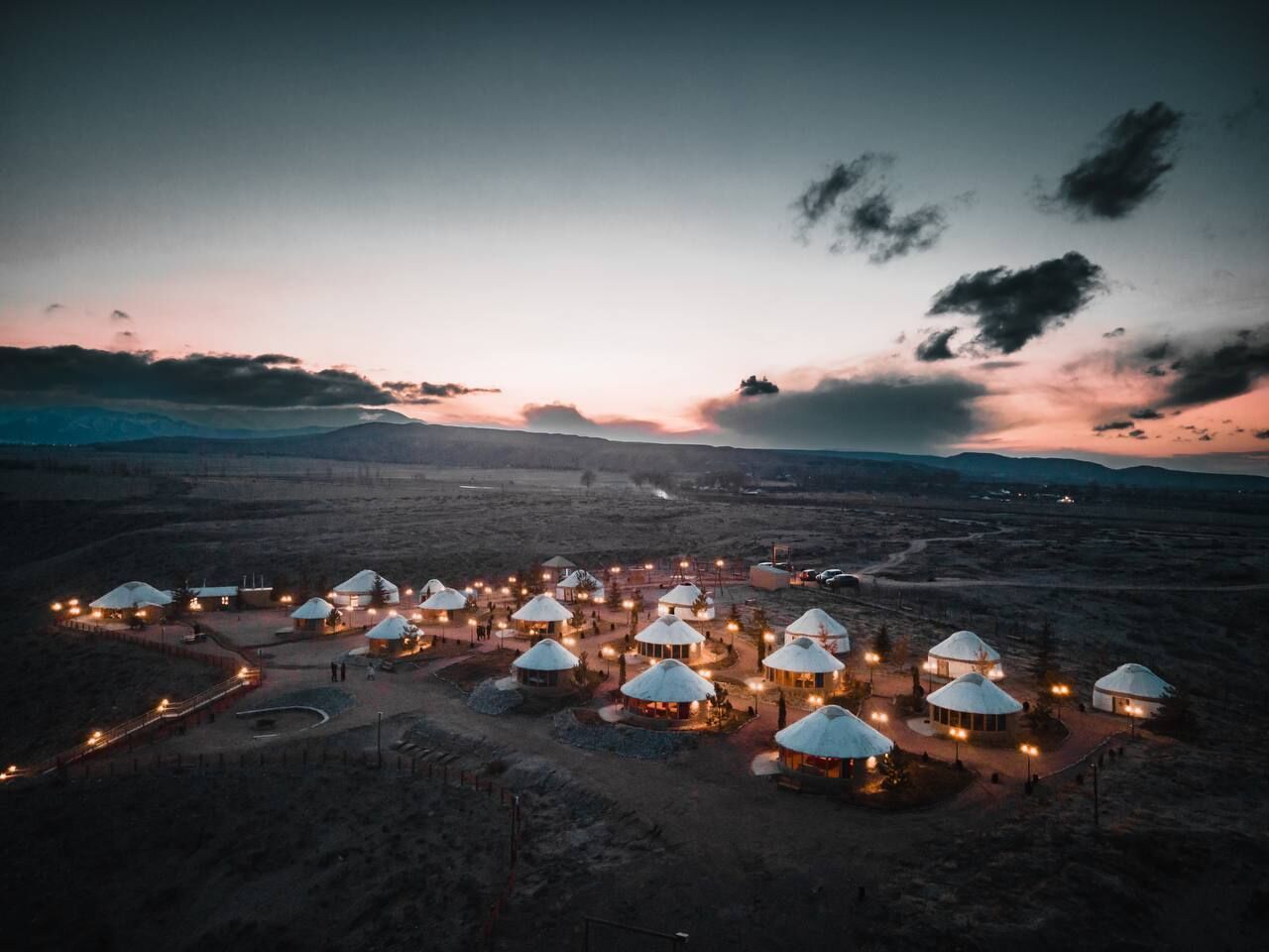 A yurt settlement with multiple yurts arranged in a circular pattern, connected by pathways, set in a vast open landscape.